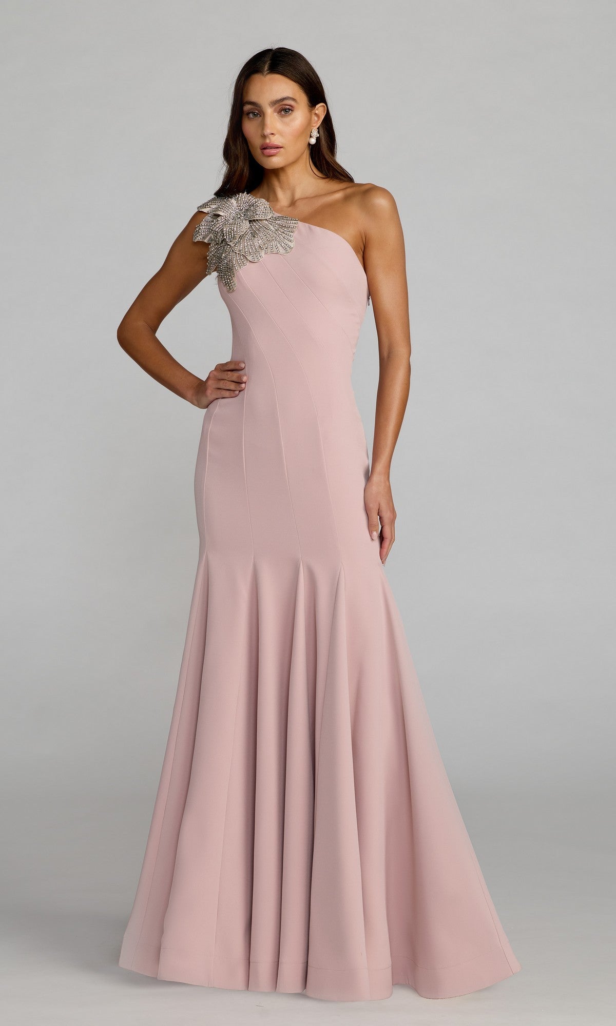 A woman stands against a plain background wearing the Mac Duggal 11810 Beaded-One-Shoulder Prom Gown in pale pink, featuring a fitted bodice and floral embellishment on the shoulder.